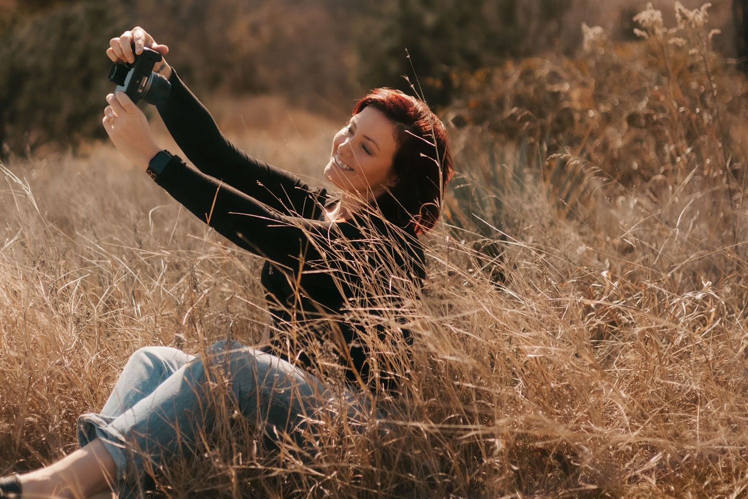 Smiling redhead woman sitting in golden dry grass field holding camera during autumn day.