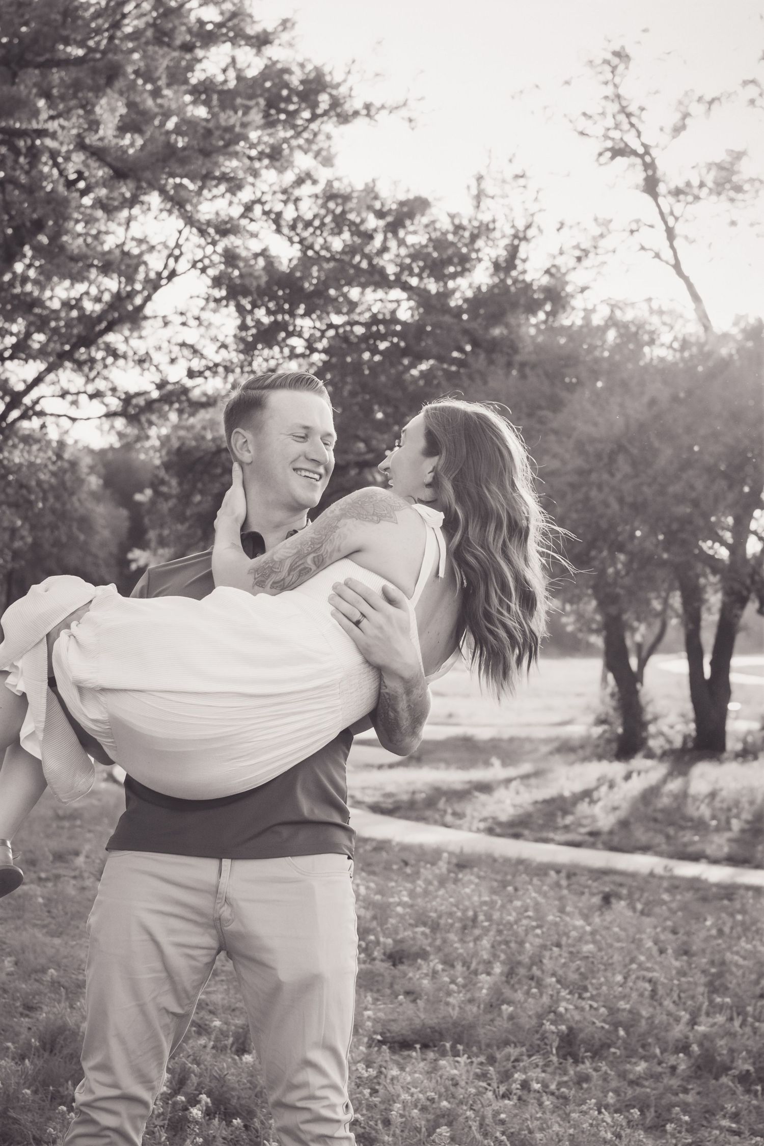 A black and white photo of a smiling man carrying a laughing woman in his arms outdoors.