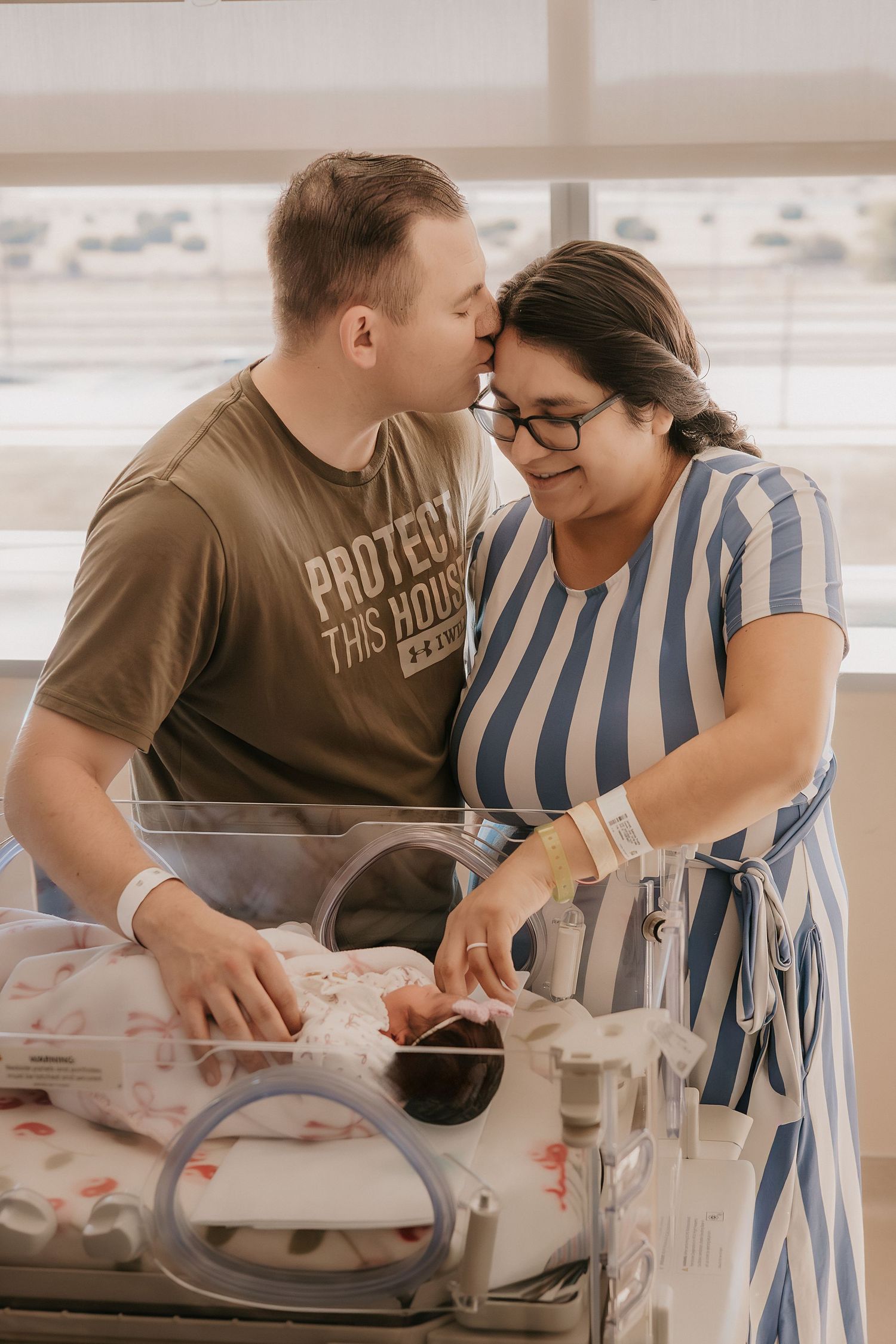 Loving couple kisses while gazing at their newborn in a hospital bassinet, wearing patient wristbands.