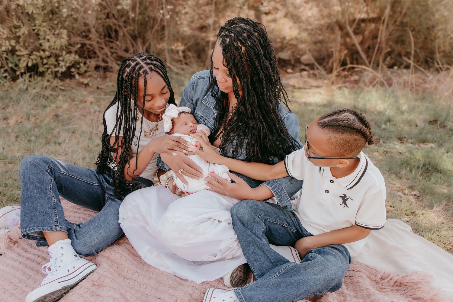 Family sitting on pink blanket outdoors with mother holding newborn baby surrounded by two children.
