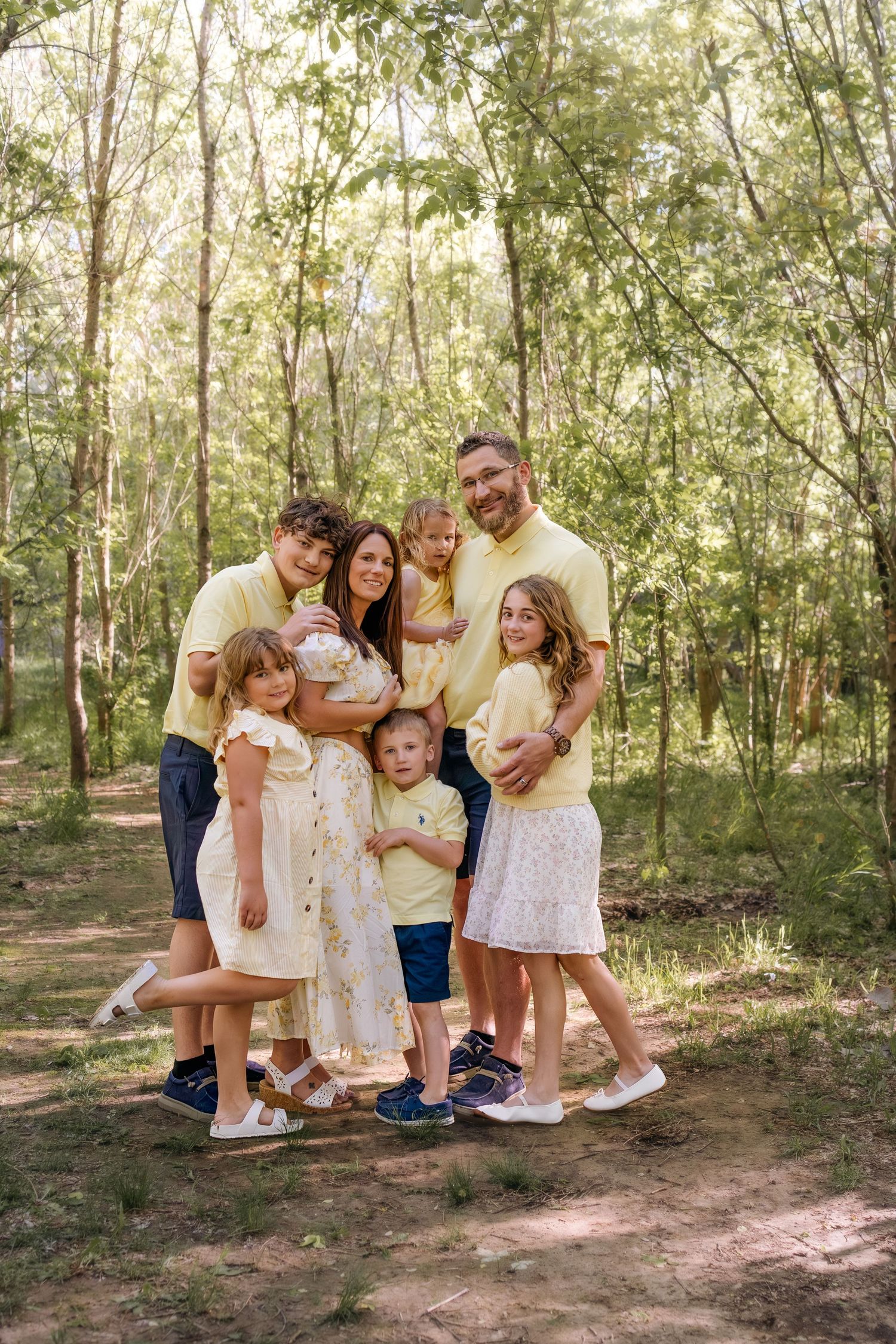 Happy family of seven posing together in a sunlit forest, wearing coordinated yellow and white outfits.