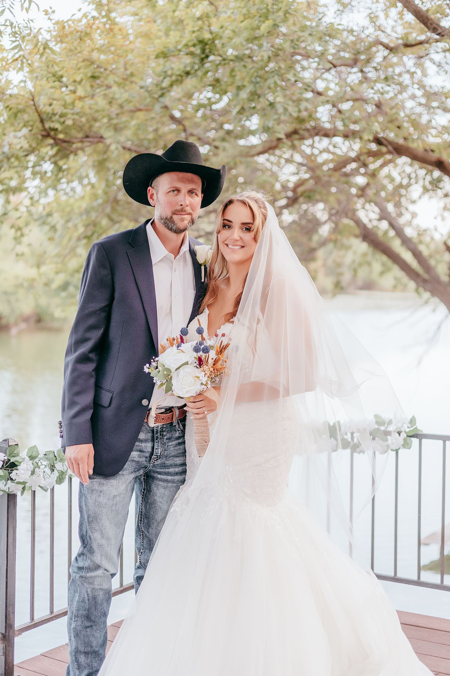 Bride in lace gown with veil holding bouquet stands beside groom in black cowboy hat and blazer on outdoor deck.