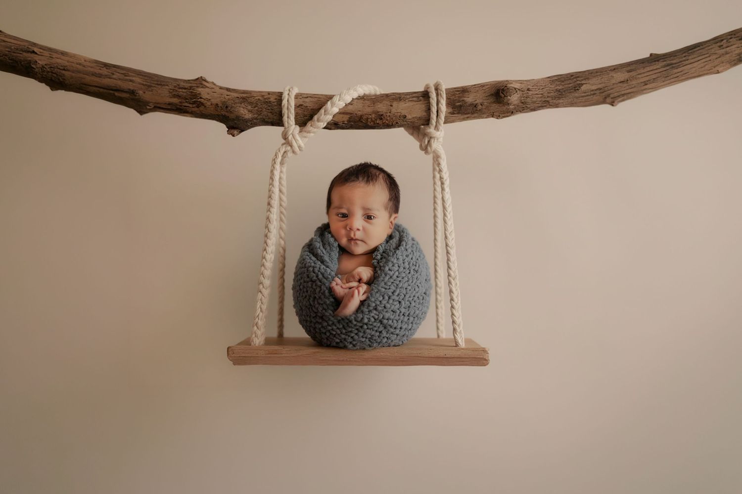Newborn baby wrapped in gray knit blanket sitting on a wooden swing suspended by rope from a rustic branch.