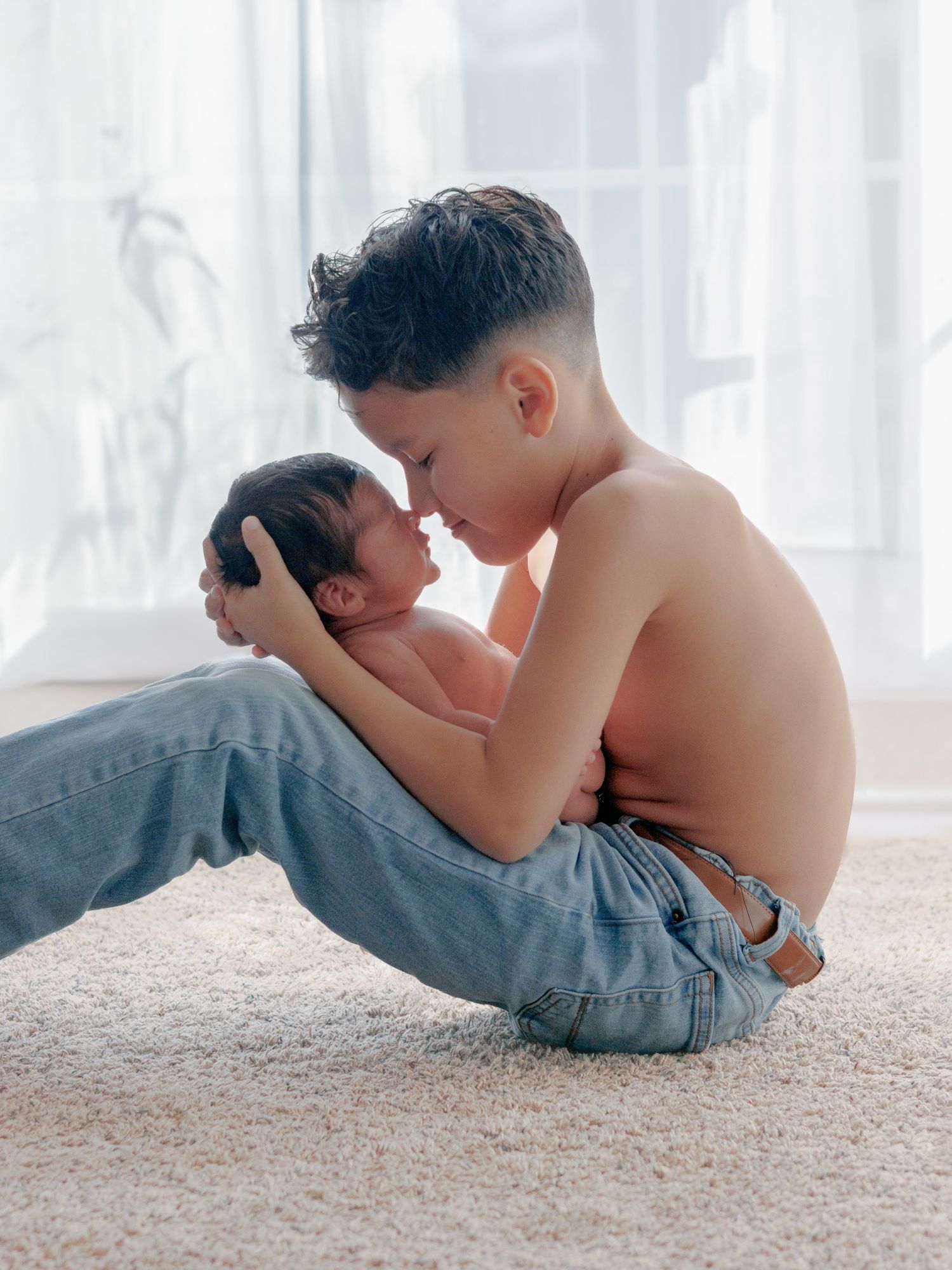 Young boy sitting on carpet holding newborn baby close, touching foreheads gently in a tender sibling moment.