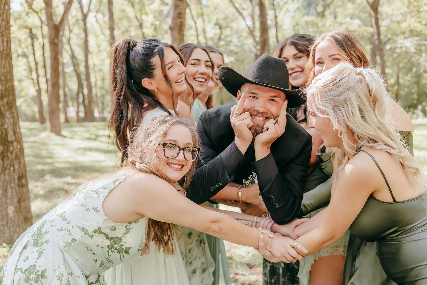 Groom in black cowboy hat surrounded by laughing bridesmaids in a sunny wooded outdoor wedding setting.