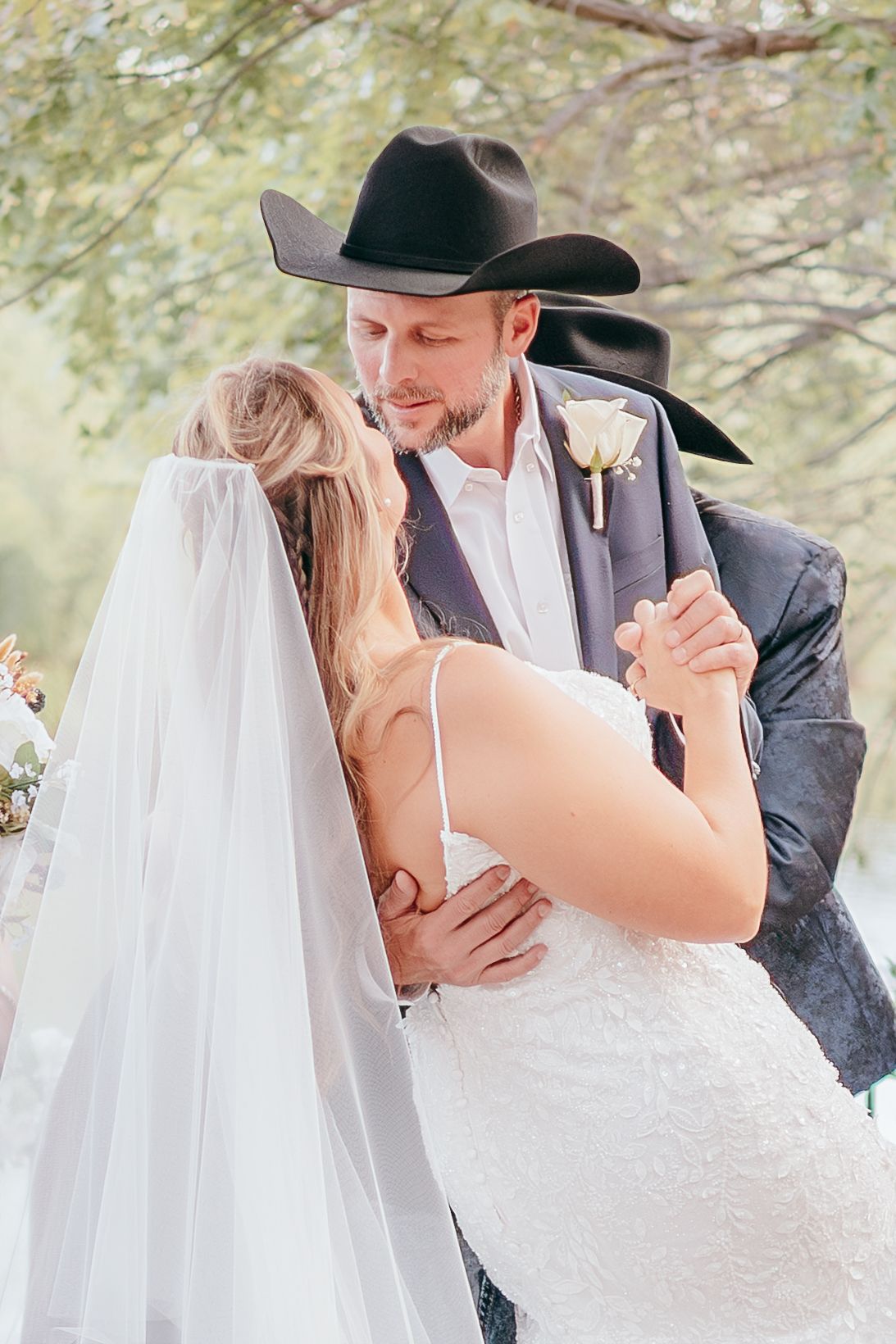 Groom in black cowboy hat embracing bride in white lace gown during first dance.