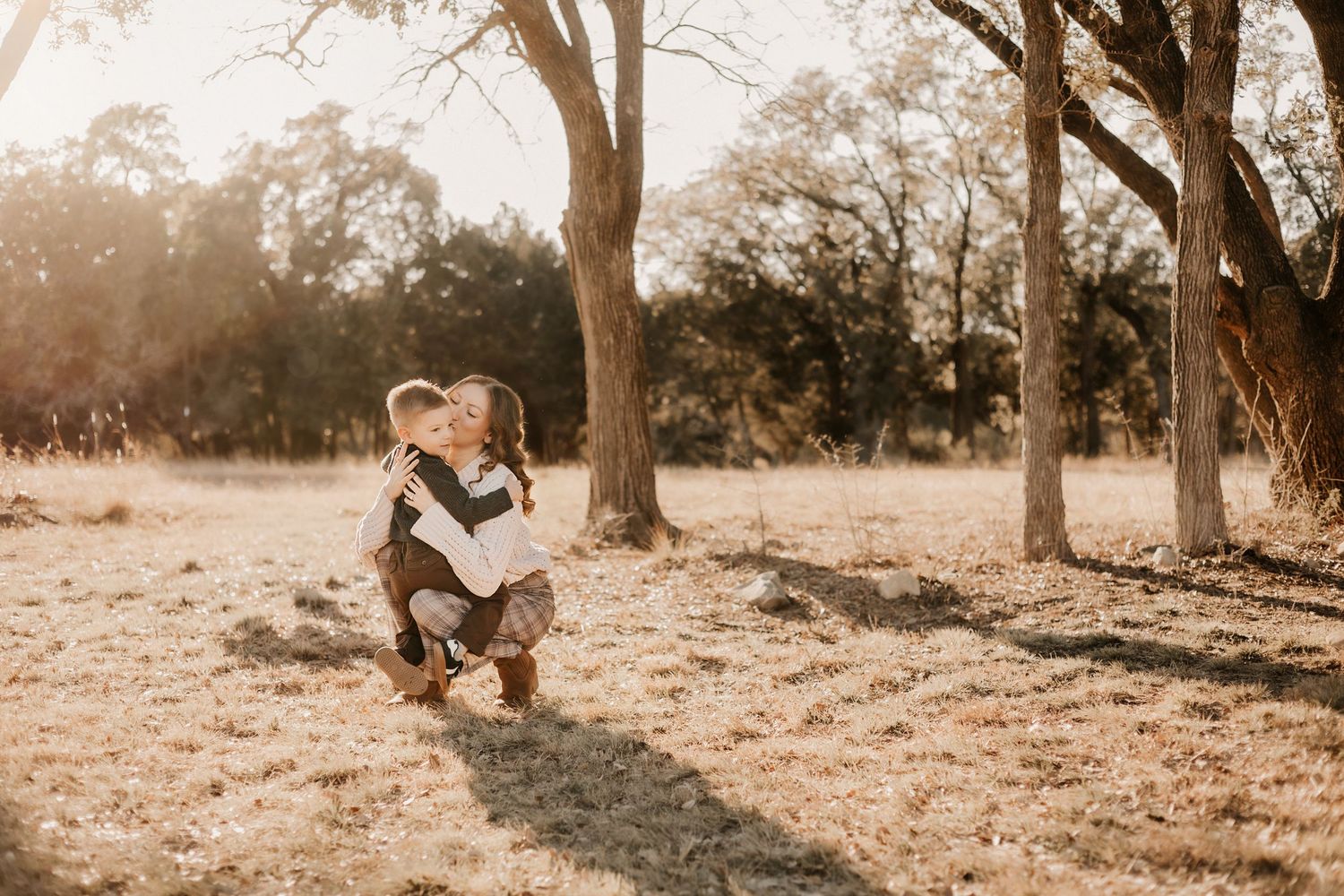 A smiling mother crouching to embrace her young son in a golden sunlit outdoor field.