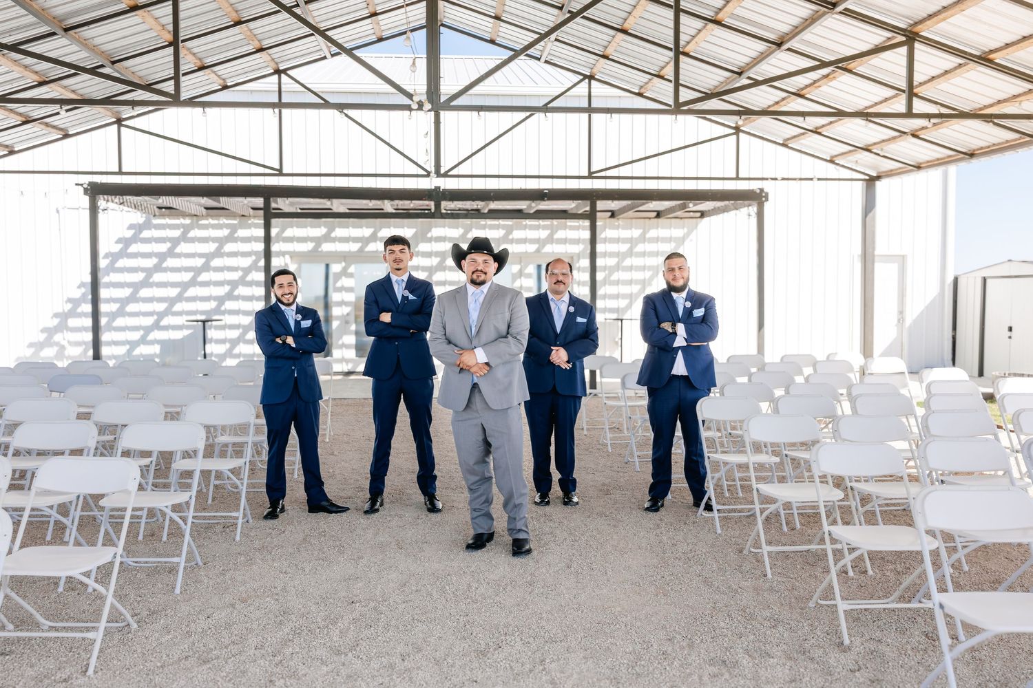 Groom in gray suit and cowboy hat poses with groomsmen in navy suits in an open air barn venue.