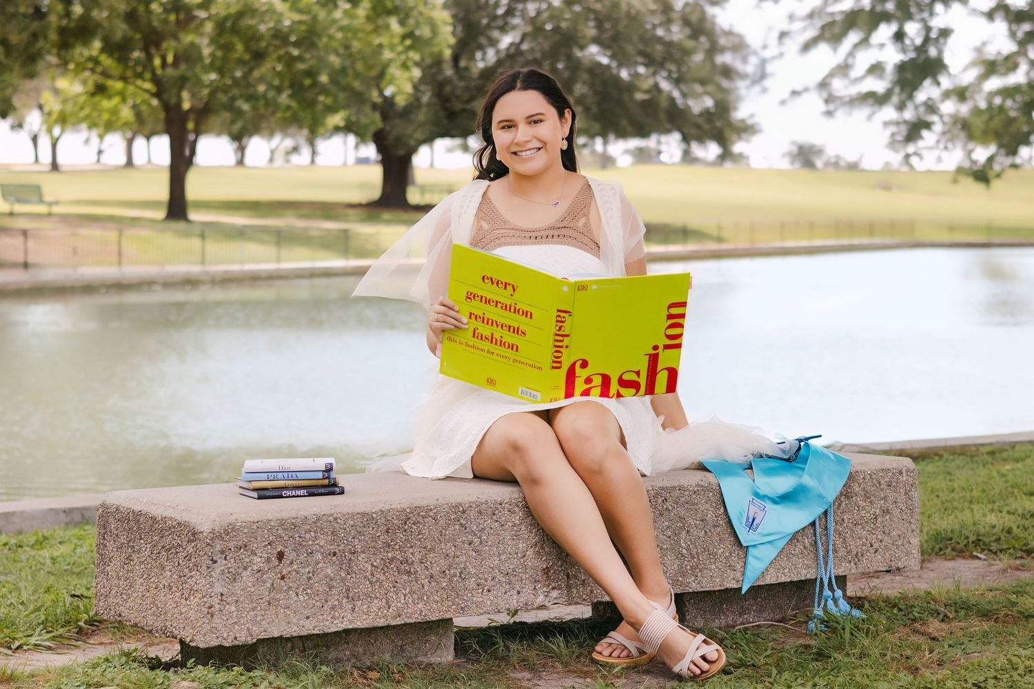 Young woman in white dress reads colorful fashion book while sitting on stone bench by pond.