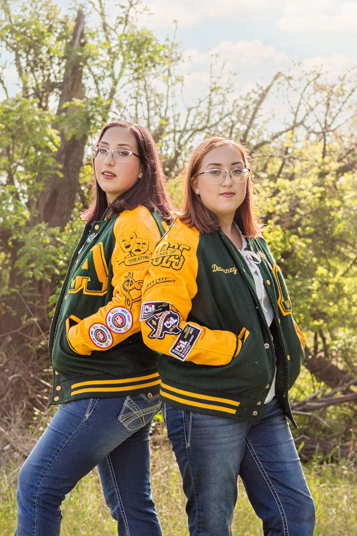 Two twin girls in matching green and yellow varsity jackets with patches pose back-to-back outdoors.