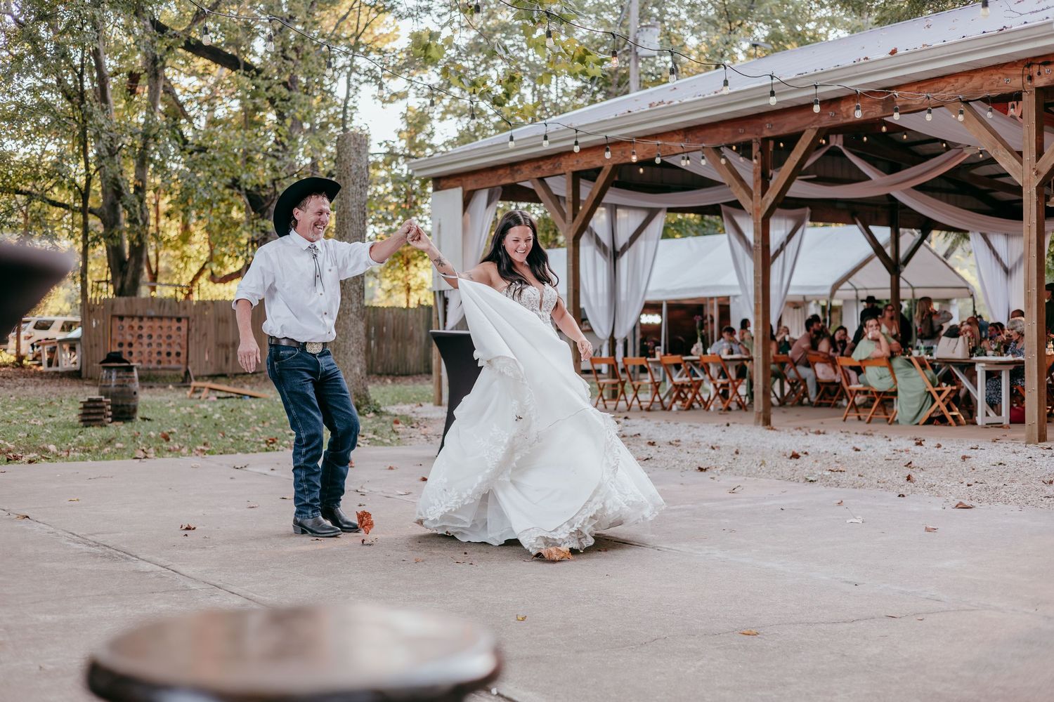 Bride in white gown and groom in cowboy hat dancing outdoors at a rustic country wedding reception.