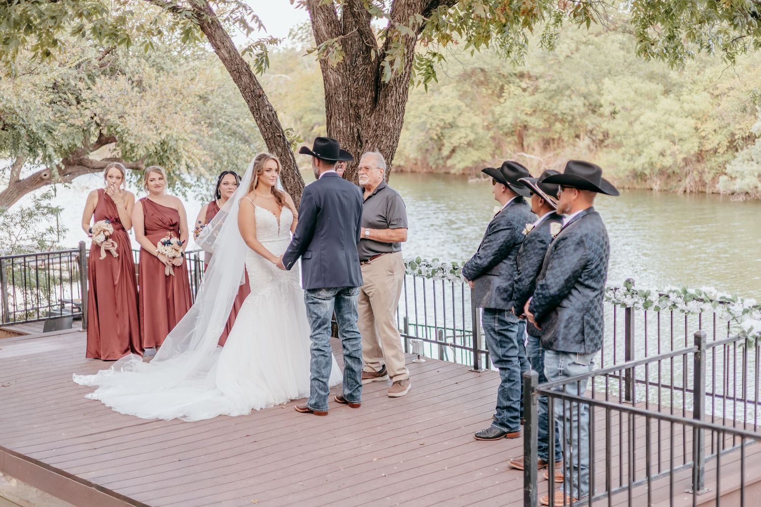 Wedding ceremony on riverside deck with bridesmaids in mauve dresses and groomsmen in cowboy hats.