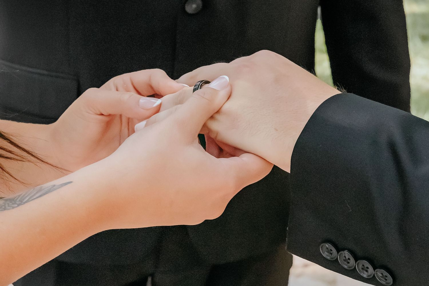Close-up of wedding ring exchange with tattooed bride's hand and groom in black suit during ceremony.