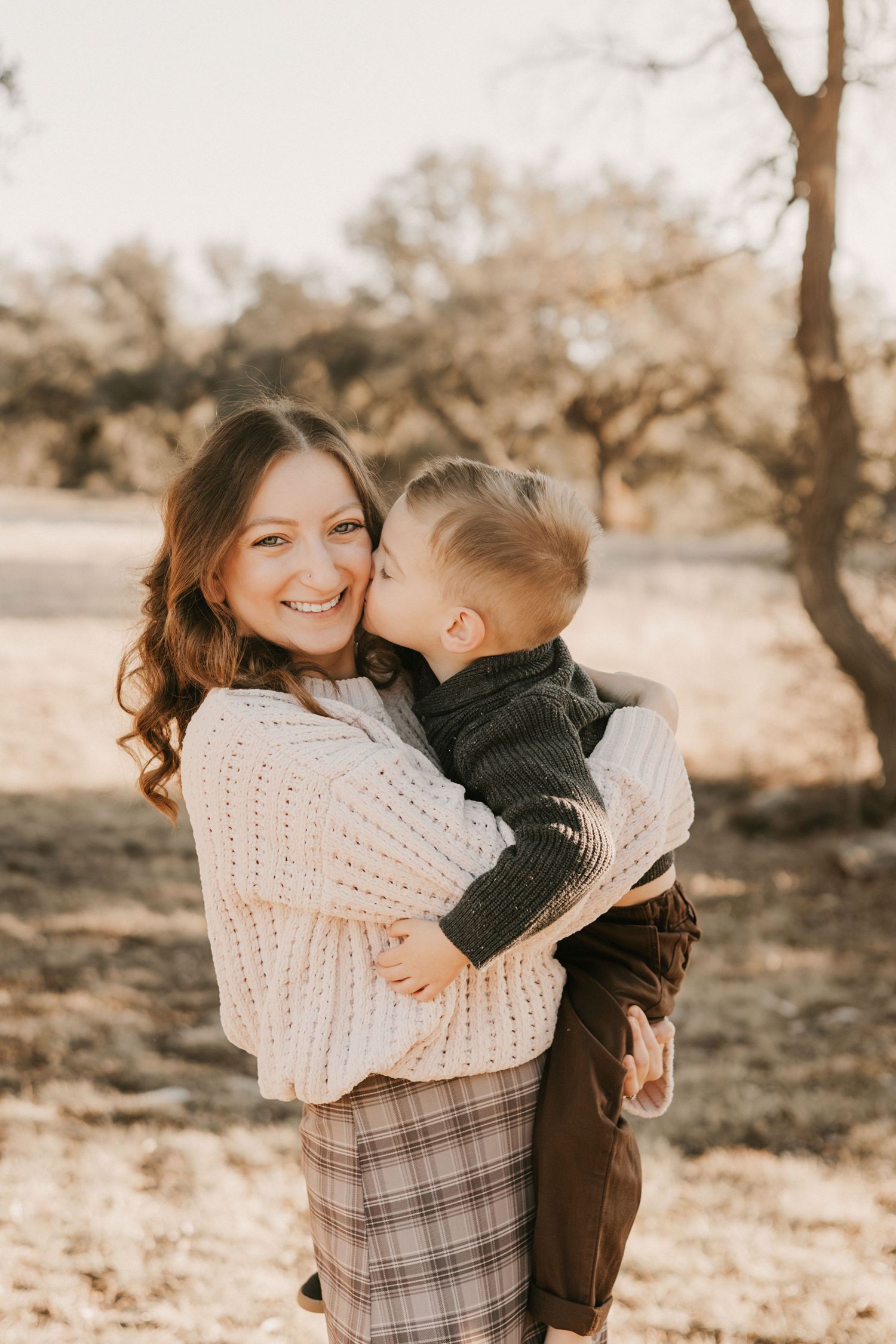 A smiling mother holds her young son as he gives her a kiss on the cheek outdoors in golden light.