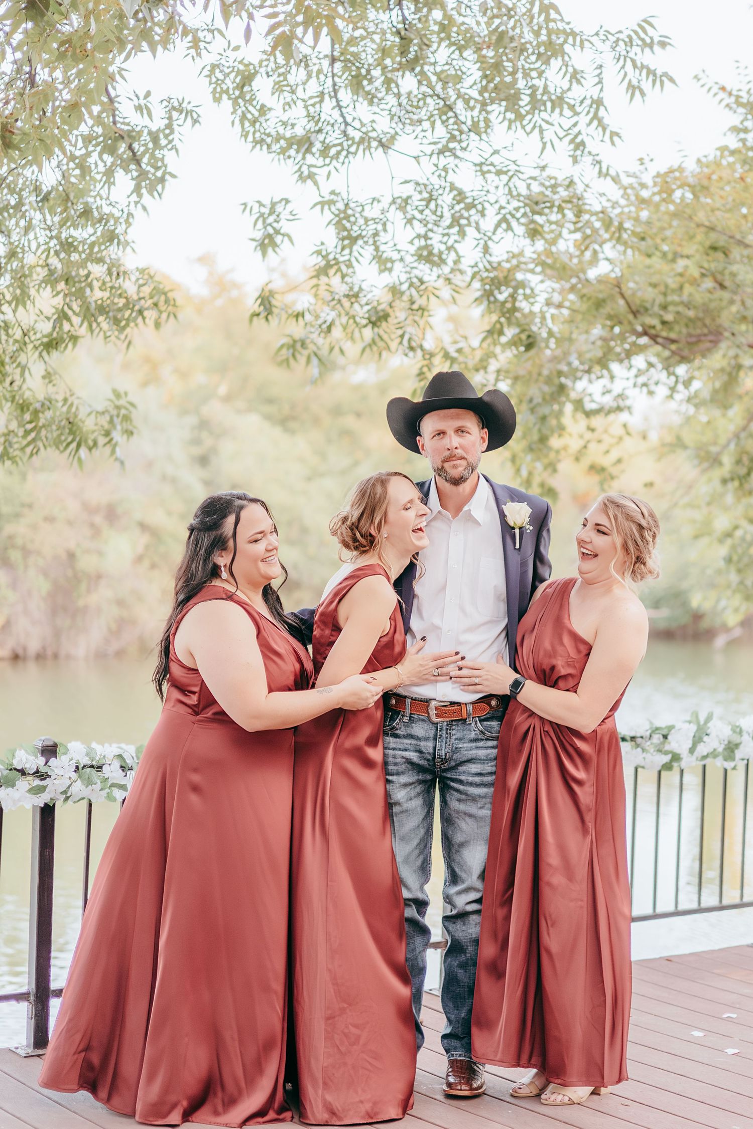Groom in black cowboy hat and jeans poses with three bridesmaids in rust-colored satin dresses on a waterfront deck.