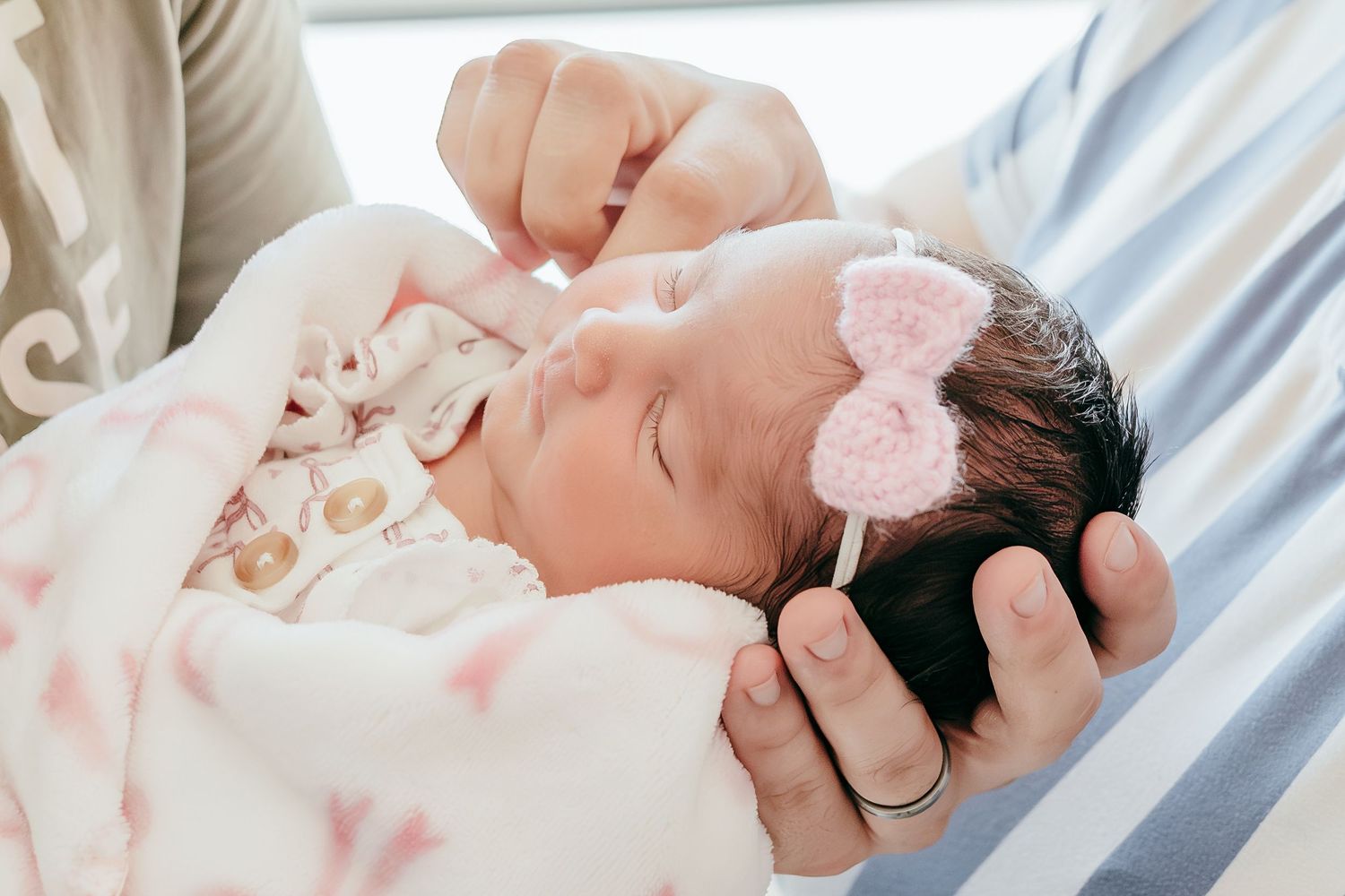Sleeping newborn baby with dark hair wearing a pink crochet bow headband cradled in parent's arms.