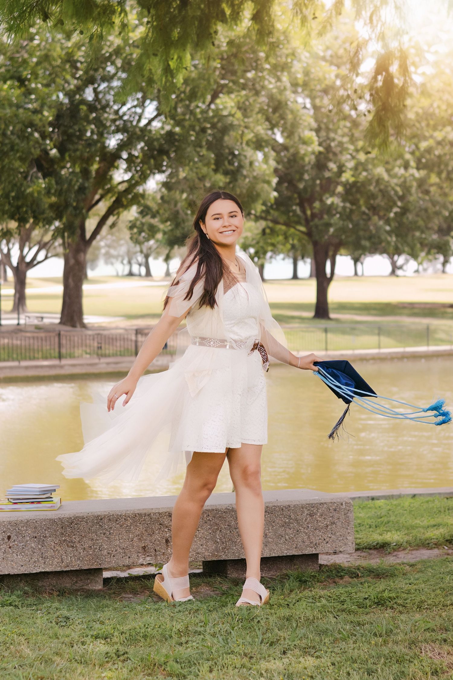 Smiling young woman in white dress twirls with blue graduation cap near sunny park pond.