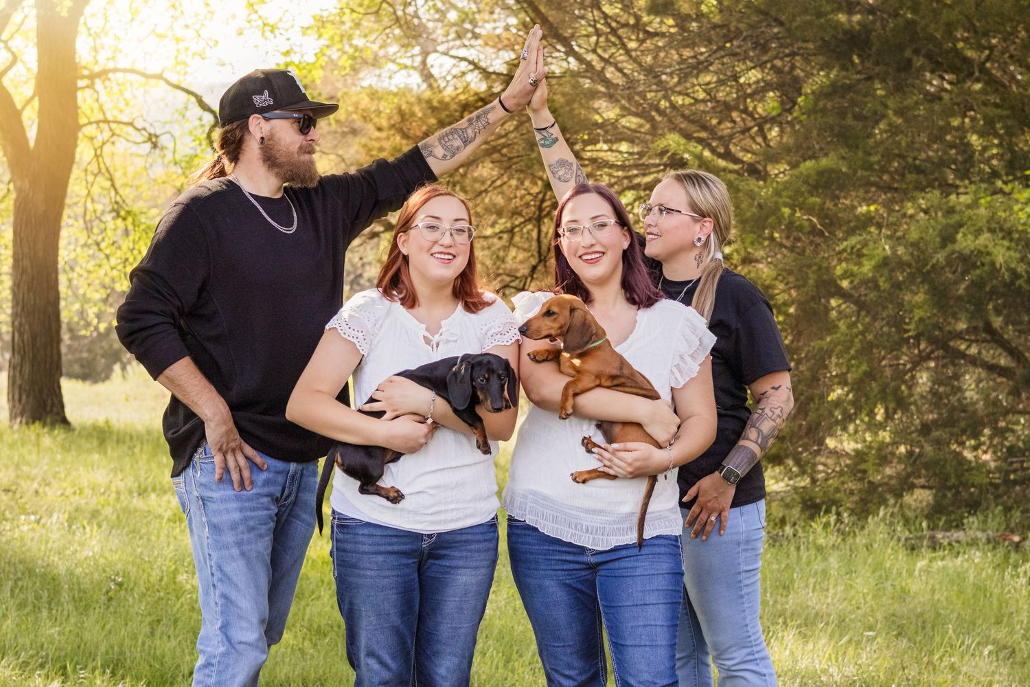 Four people smiling outdoors in sunlight, two holding dachshund puppies, posing together in a green park setting.