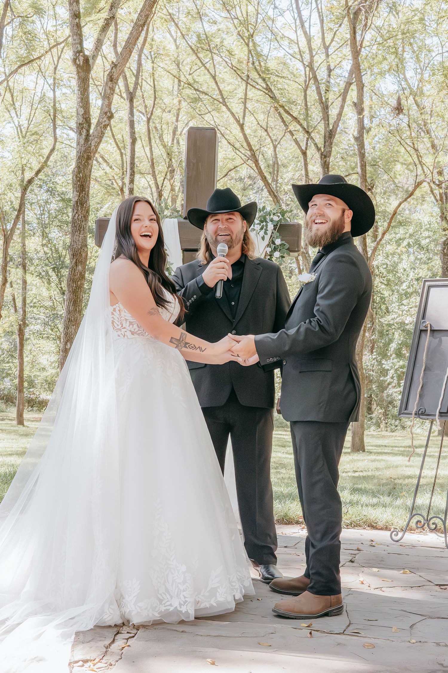 Bride and groom laugh joyfully during outdoor wedding ceremony with officiant holding microphone in wooded setting.