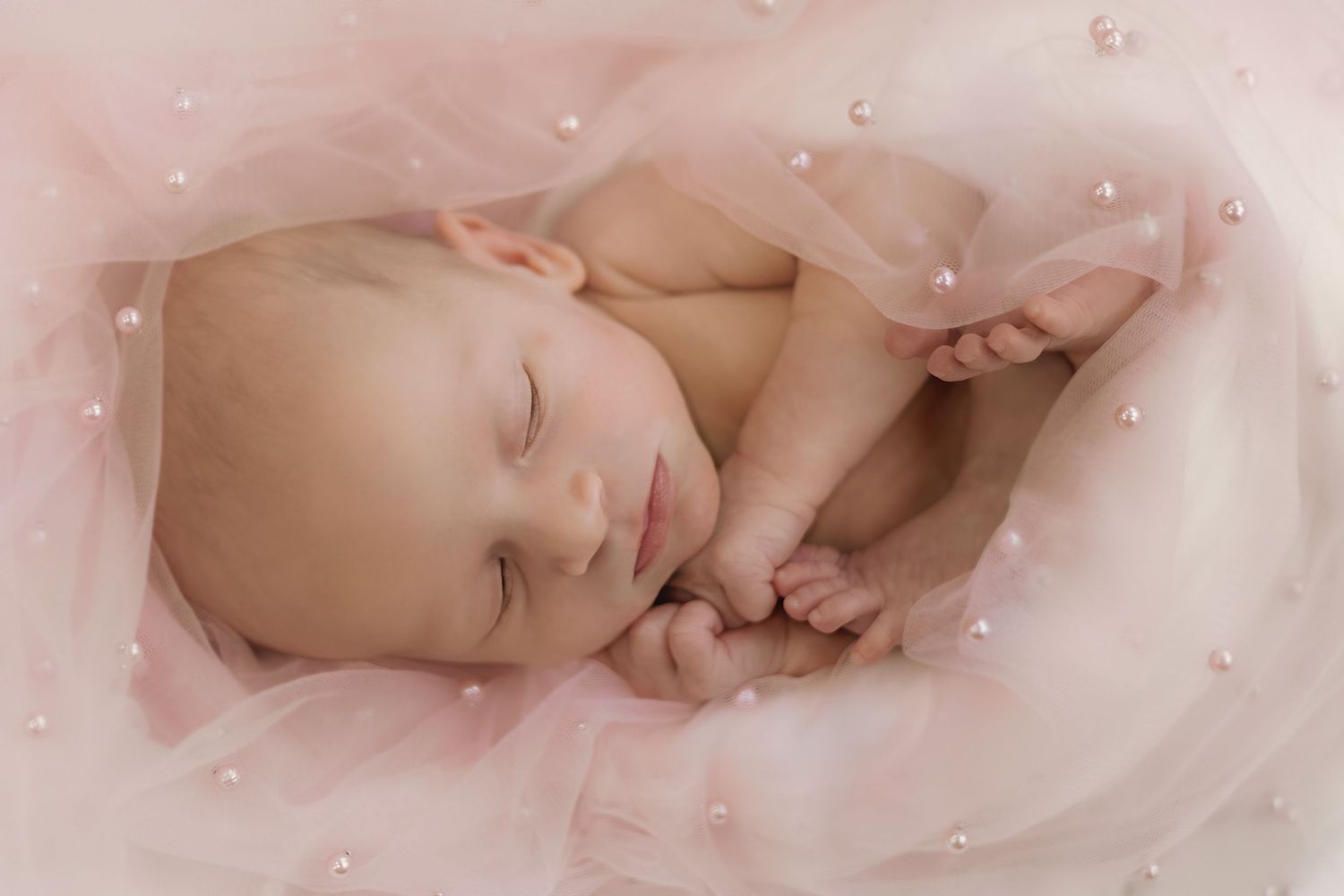 Serene newborn baby resting peacefully on delicate pink tulle with pearl accents.