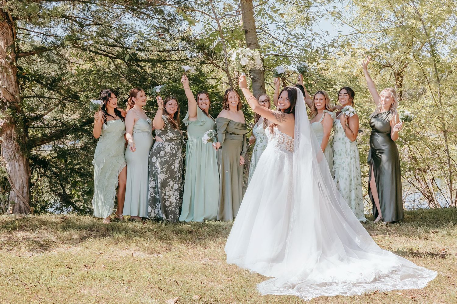 Bride and bridesmaids in sage green dresses celebrate outdoors raising bouquets under trees on sunny wedding day.