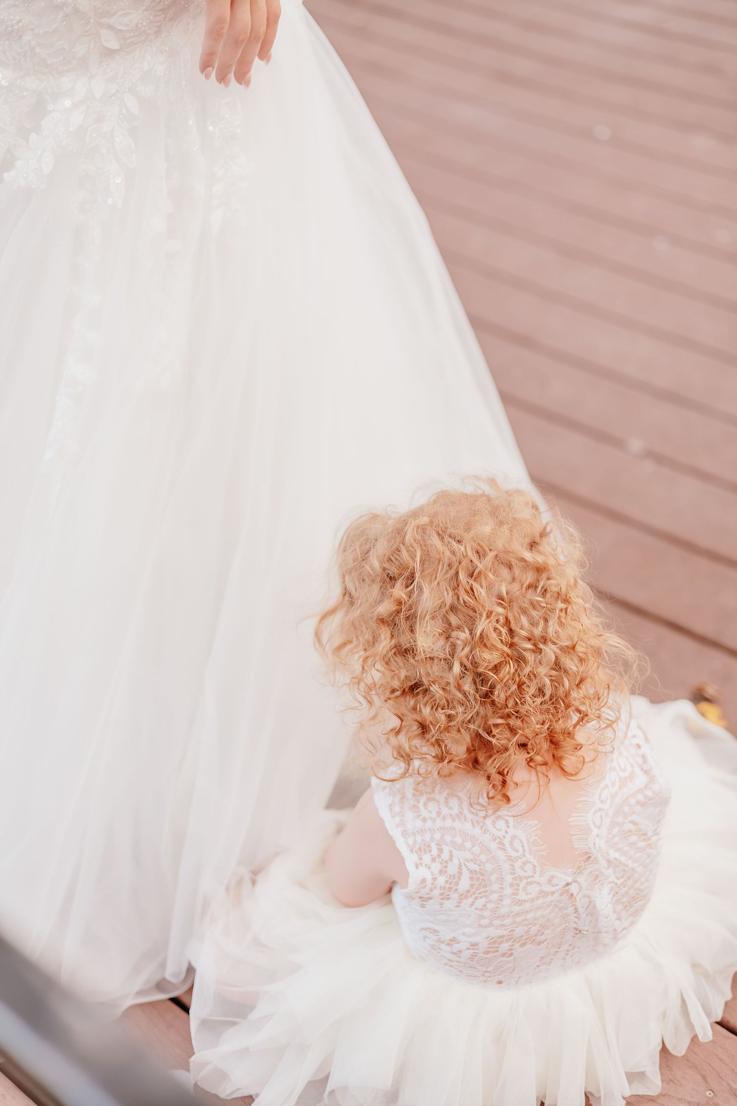 Curly-haired flower girl in white lace dress sitting beside bride's wedding gown.