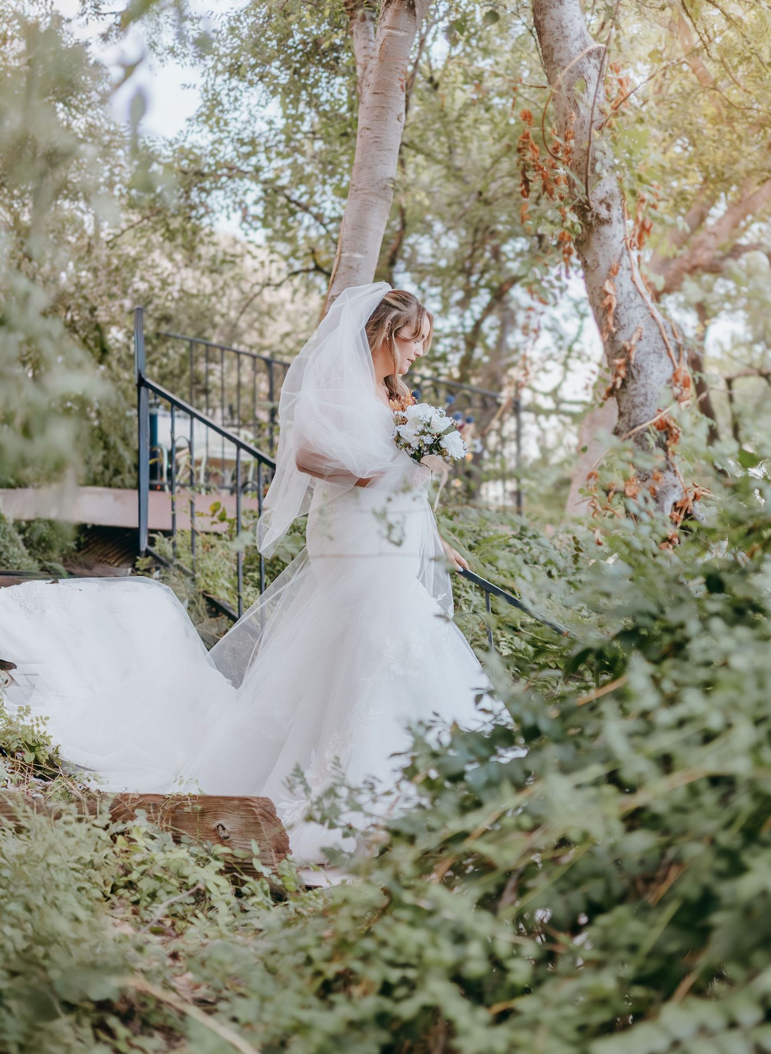 Bride in flowing white gown and veil walking among lush green trees and foliage outdoors.