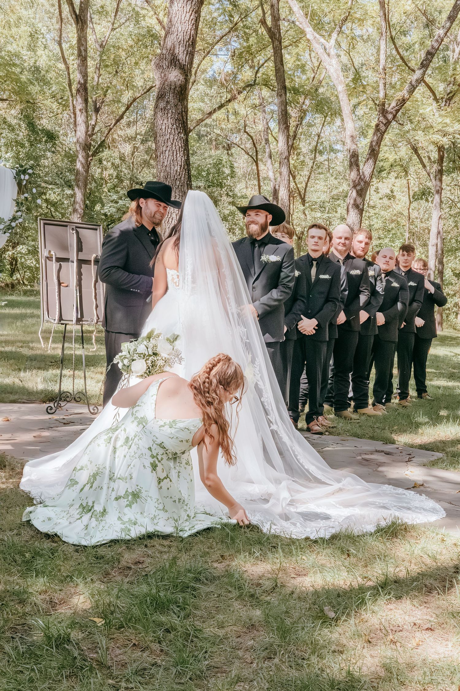 Outdoor wedding ceremony with bride in floral gown, groom in black cowboy hat, and groomsmen lined up in wooded setting.