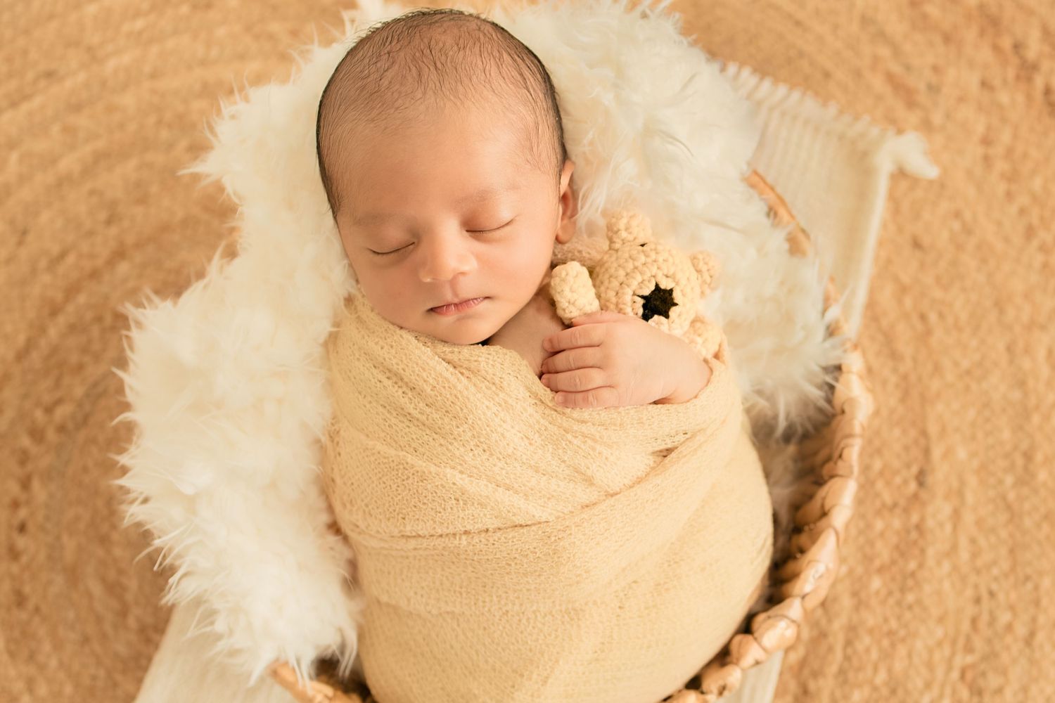 Newborn baby swaddled in yellow wrap sleeping in basket with white fur and teddy bear.