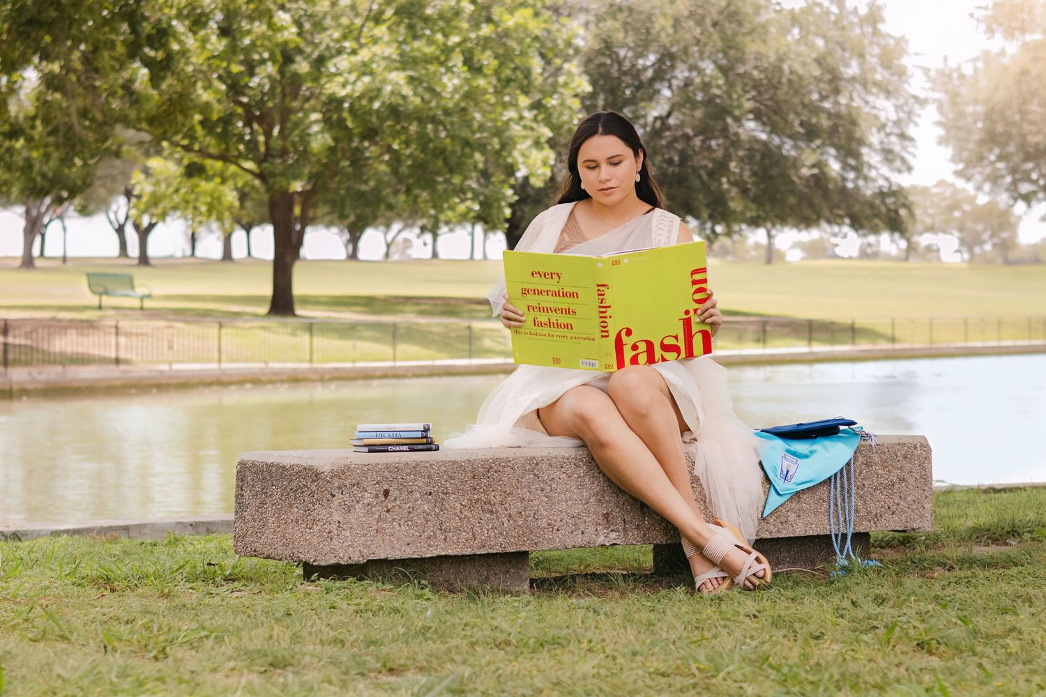 Senior Girl sitting on stone bench in park reading a yellow fashion book near a pond on a sunny day.