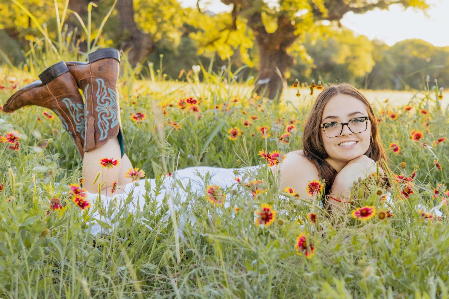 Young woman in glasses lying in a wildflower field wearing cowboy boots, smiling outdoors in golden sunlight.