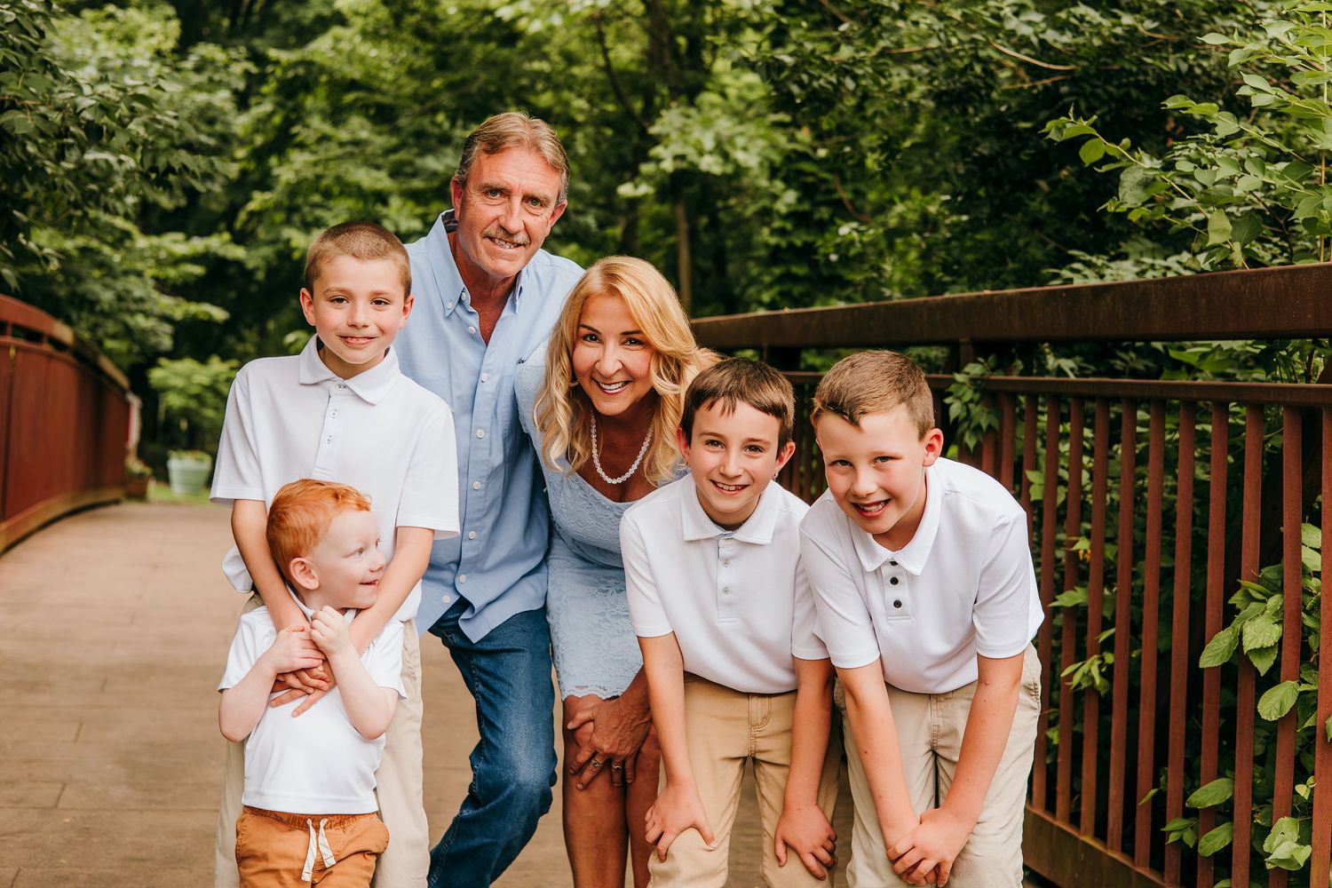 Family of six smiling on a wooden bridge surrounded by lush green trees during an outdoor portrait session.