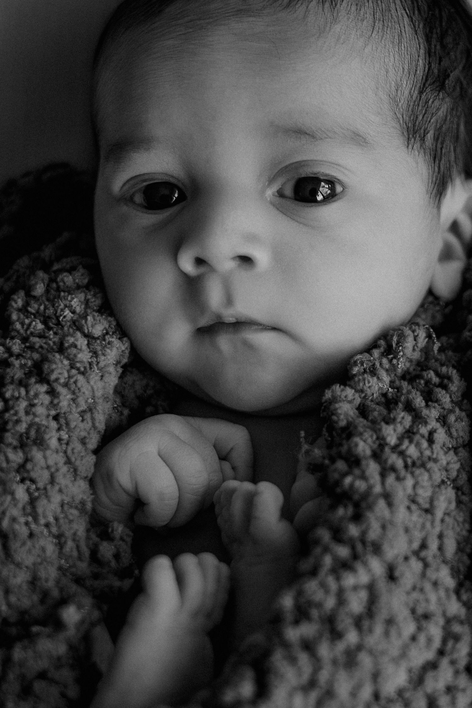 Black and white close-up portrait of an alert newborn baby wrapped in a textured knit blanket.
