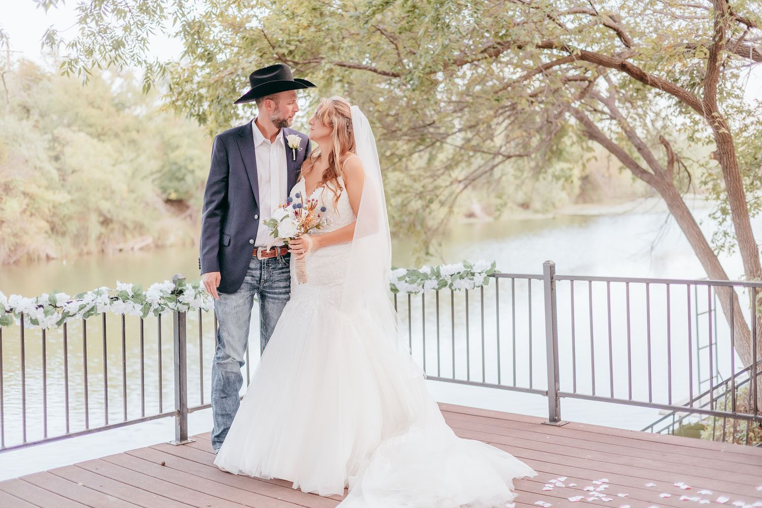Bride and groom in cowboy hat sharing romantic moment on deck near river.