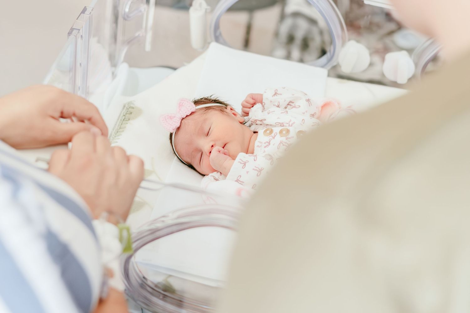 Newborn baby girl with pink headband sleeping peacefully in hospital bassinet while parents watch over her.
