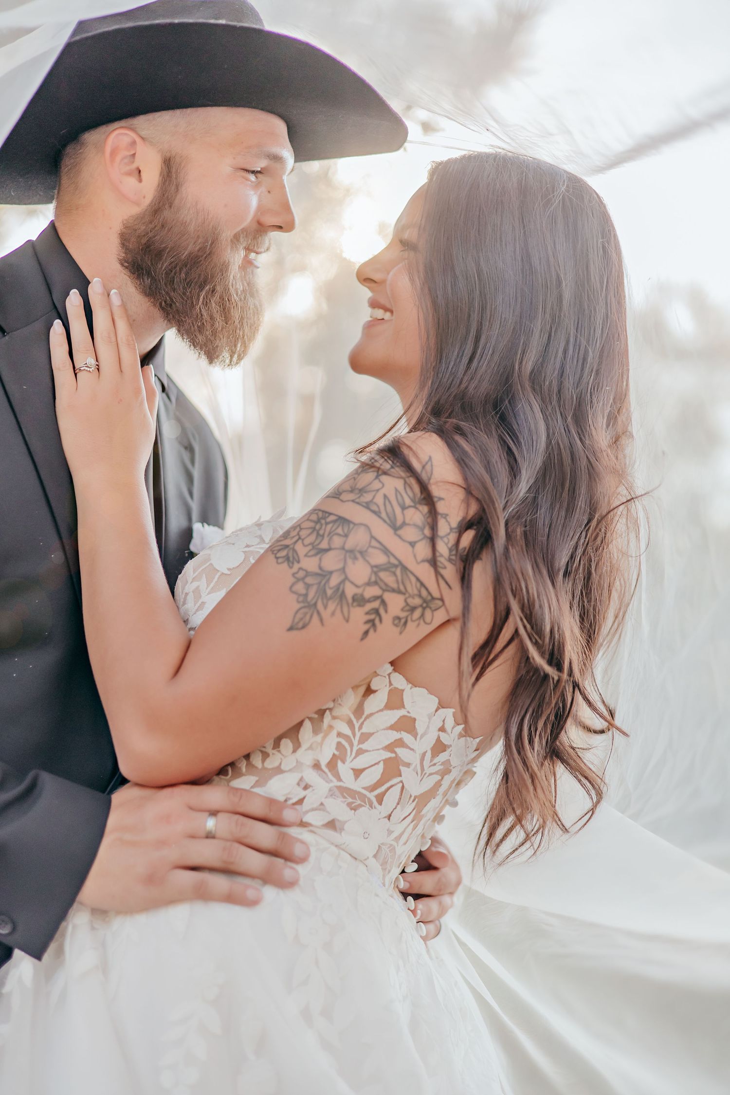 Bride and groom sharing romantic moment under veil, groom in black cowboy hat, bride with floral tattoo.