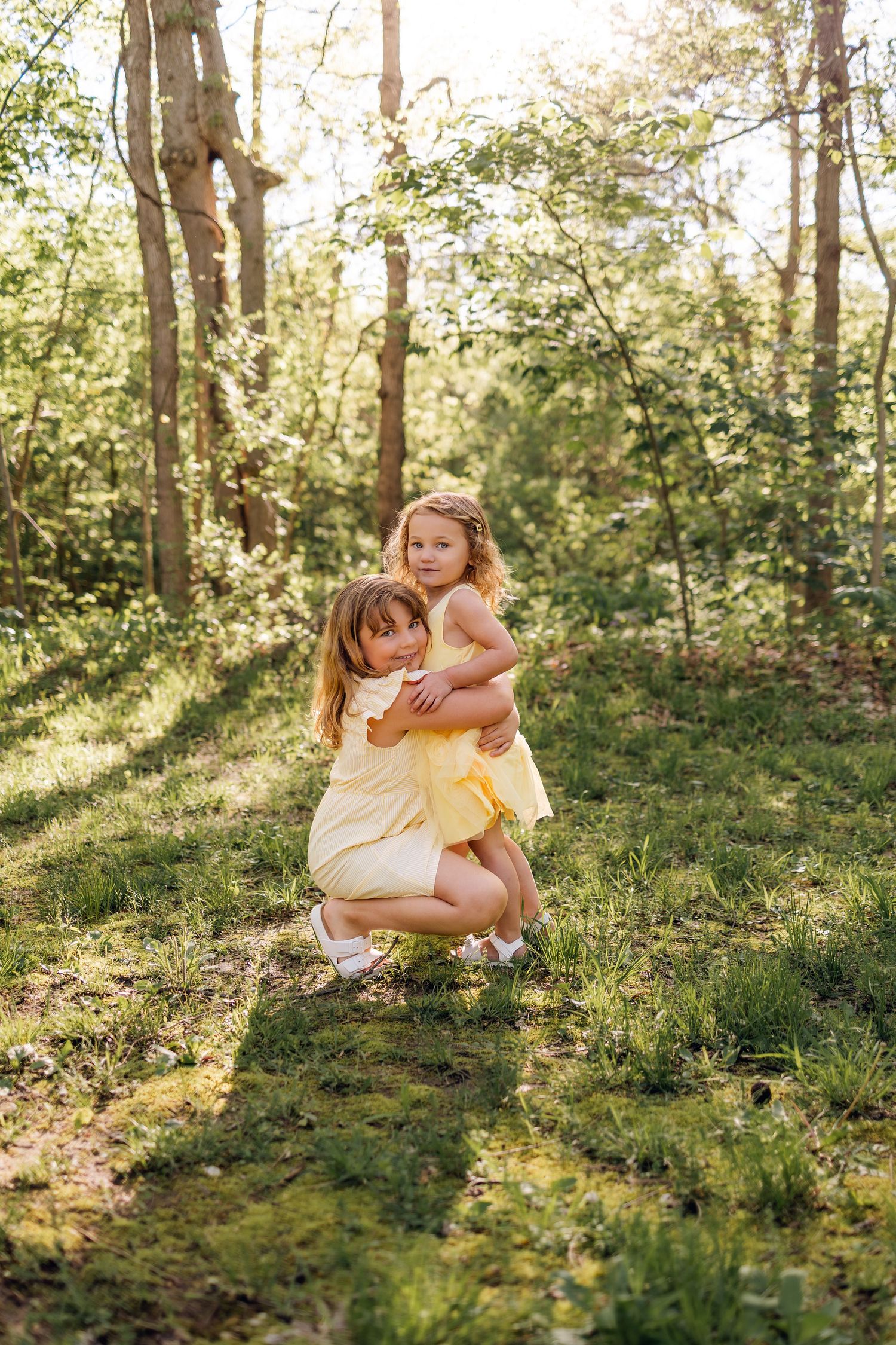 Two young girls in matching yellow dresses share a warm hug in a sunlit green forest clearing.