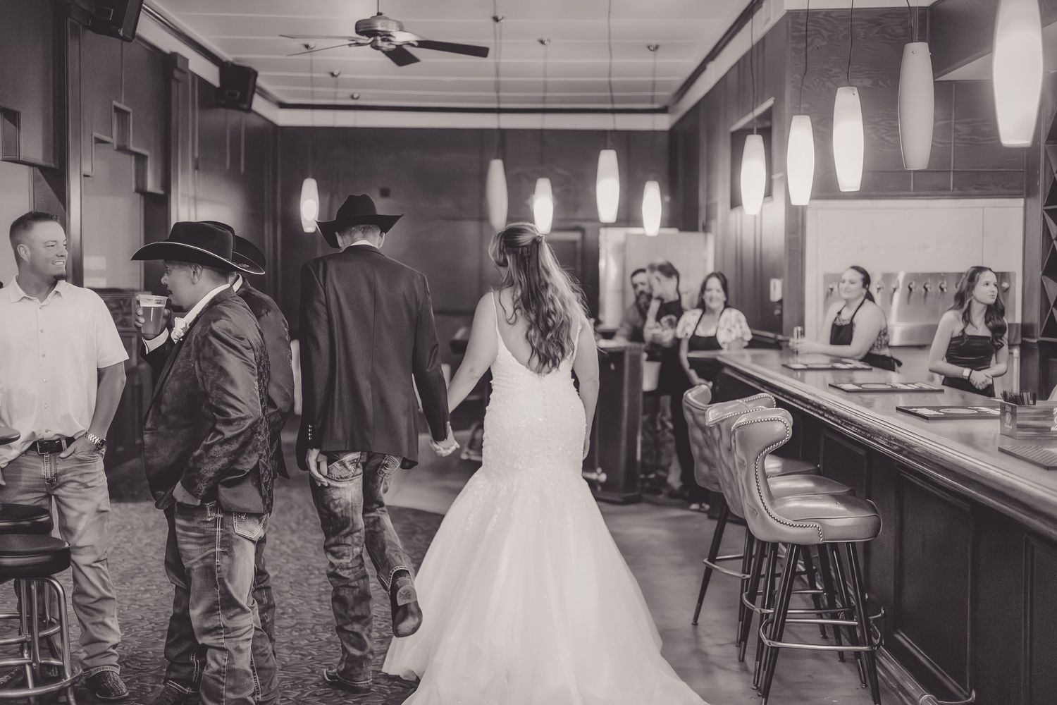 Black and white photo of a bride in a wedding gown walking through an elegant bar venue with guests nearby.