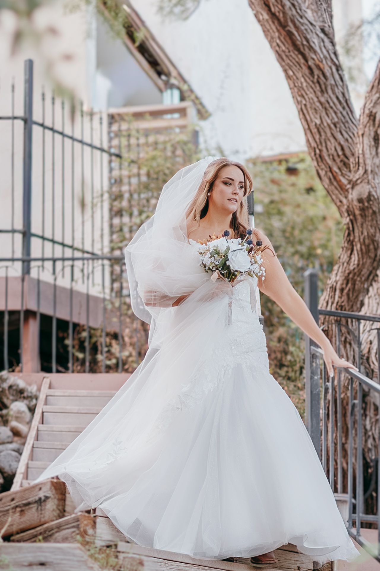 Bride twirling in fitted white gown with veil and bouquet on wooden steps outdoors.