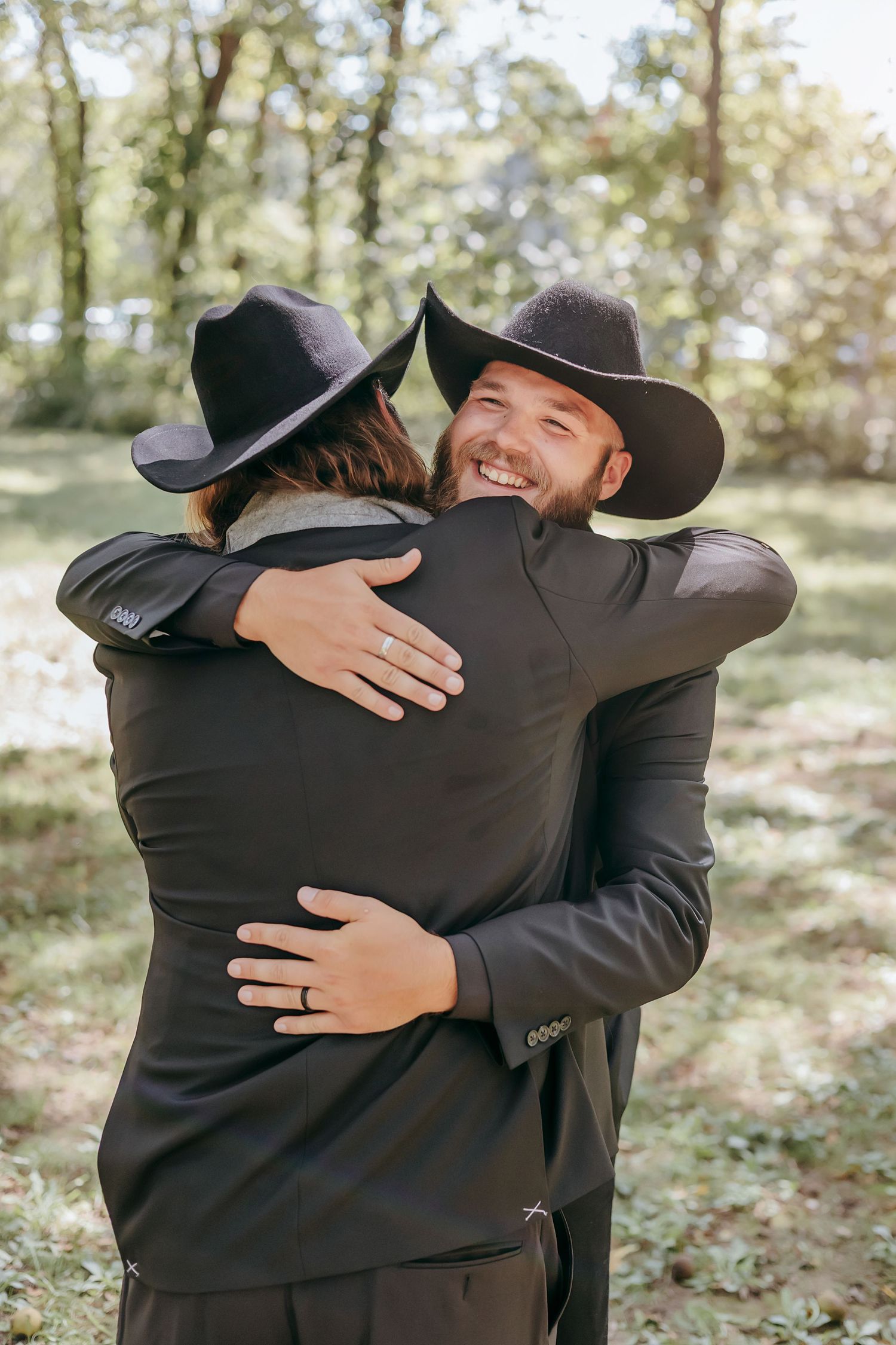 Two grooms in black suits and cowboy hats sharing a warm embrace and smiling in outdoor wooded setting.