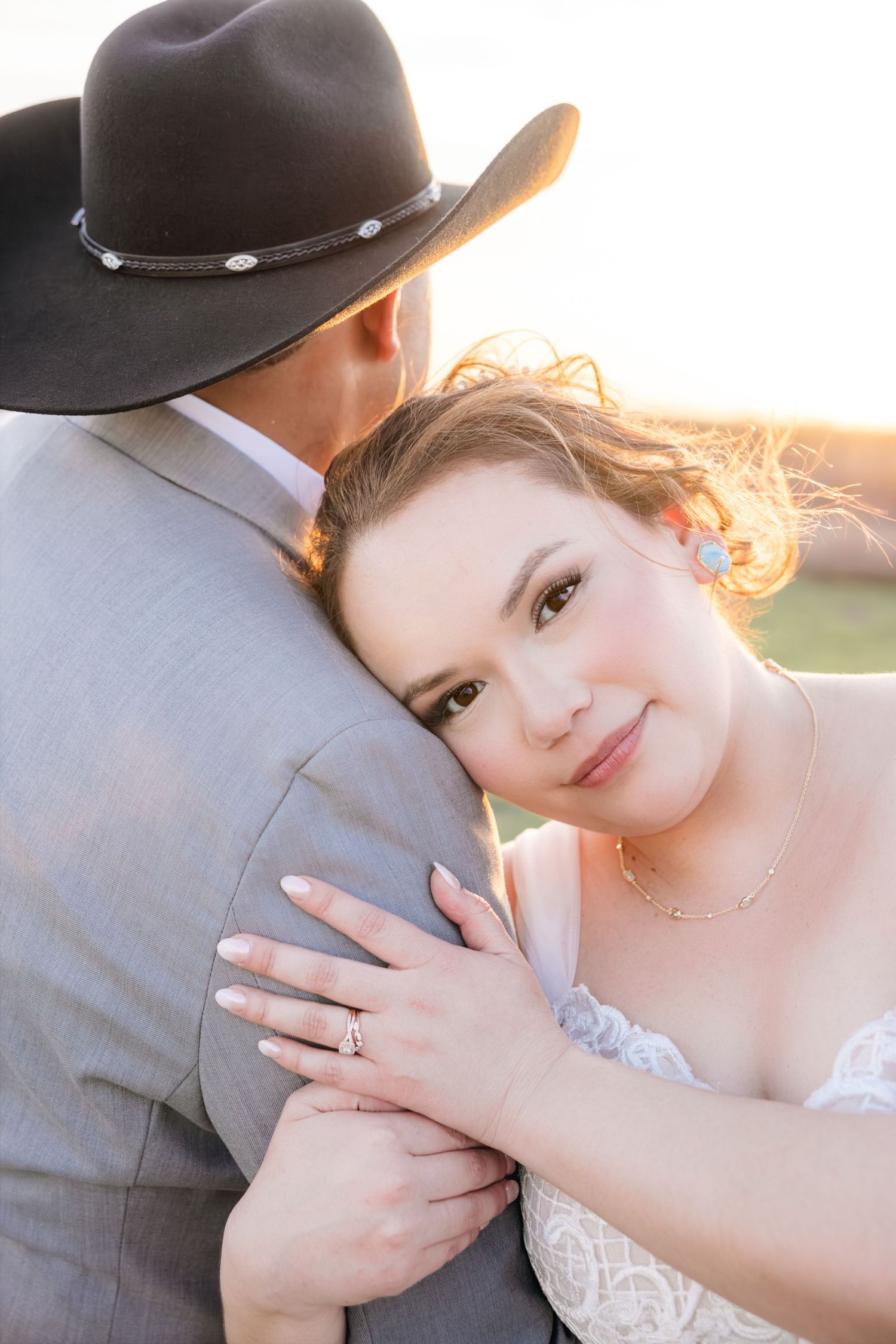 Bride leans on groom's shoulder displaying wedding ring as golden sunset light glows behind them.