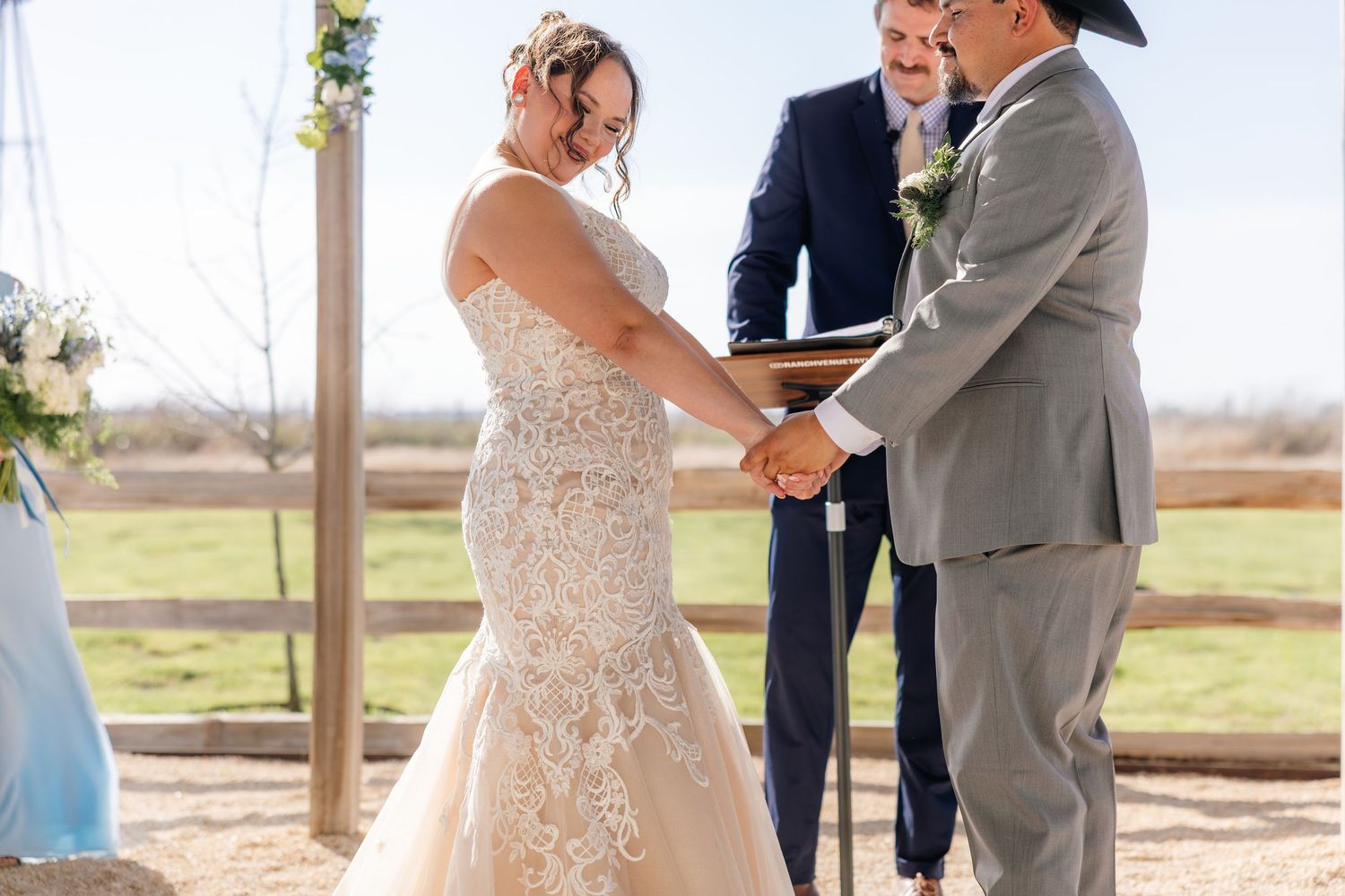 Bride and groom hold hands during outdoor rustic wedding ceremony with officiant and guests in background.
