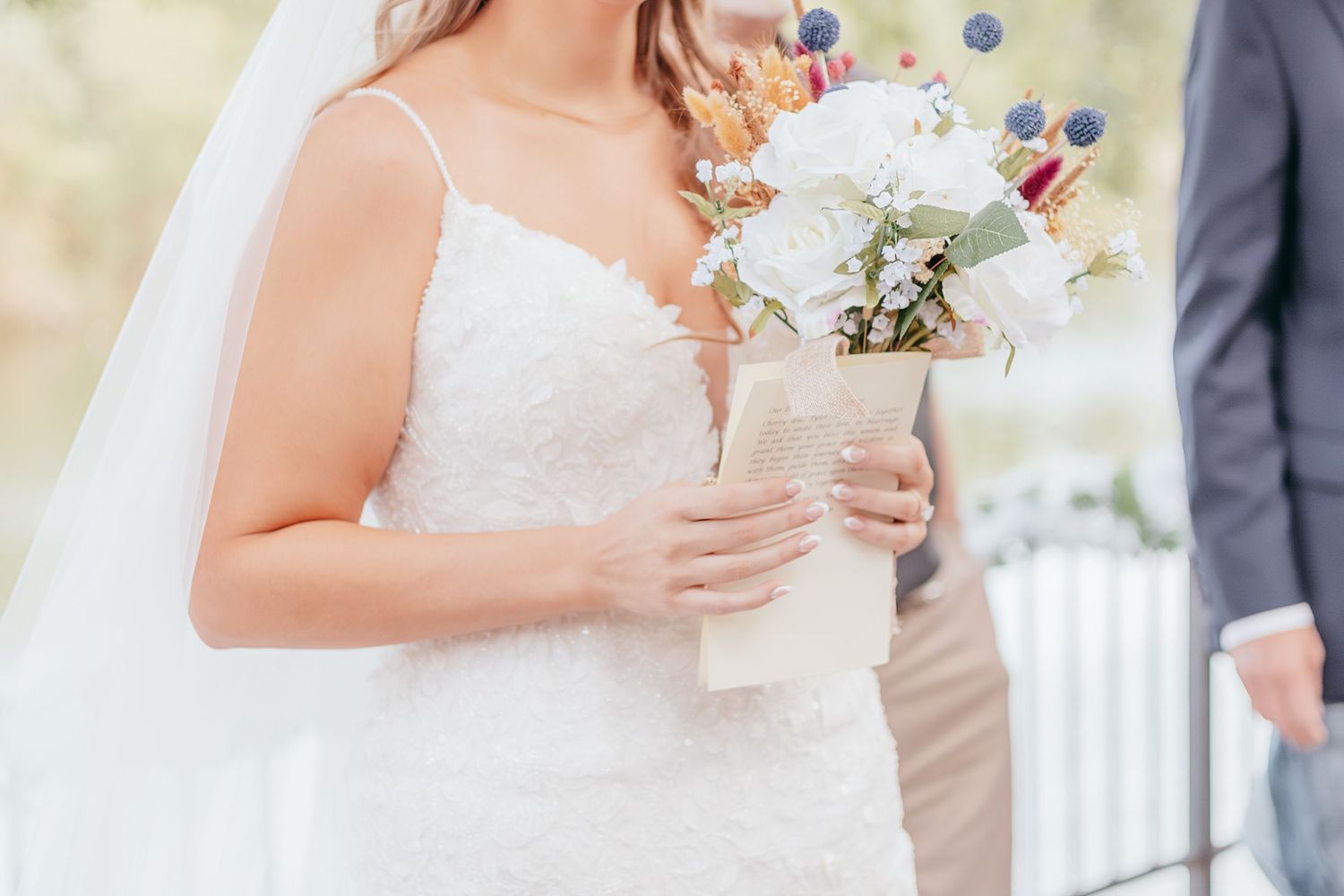 Bride in lace wedding gown holding white floral bouquet with eucalyptus and dried flowers at outdoor ceremony.