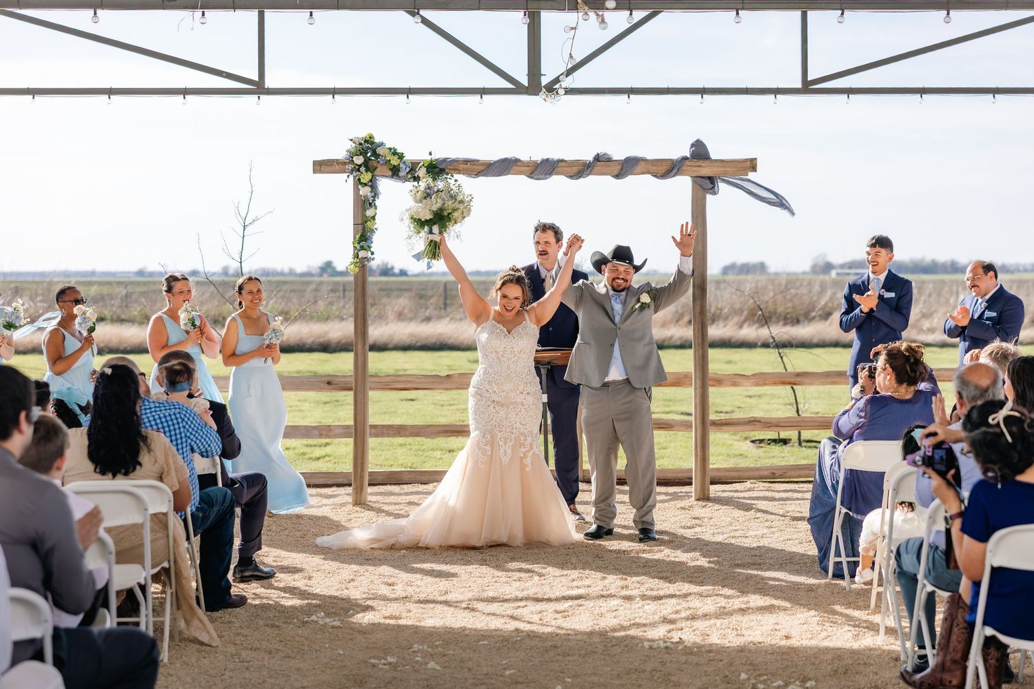 Joyful bride and groom celebrate at rustic outdoor wedding ceremony with bridal party and guests applauding under wooden arch.