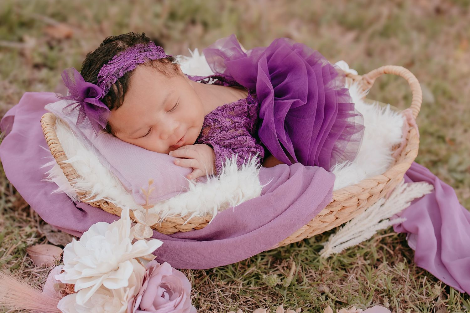 Sleeping newborn baby in purple lace dress and floral headband nestled in wicker basket with white fur.