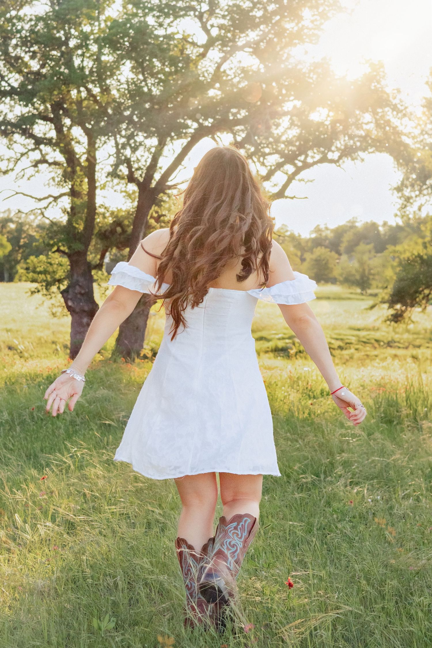 Woman in white off-shoulder dress and cowboy boots walking through sunny green meadow with trees in background.