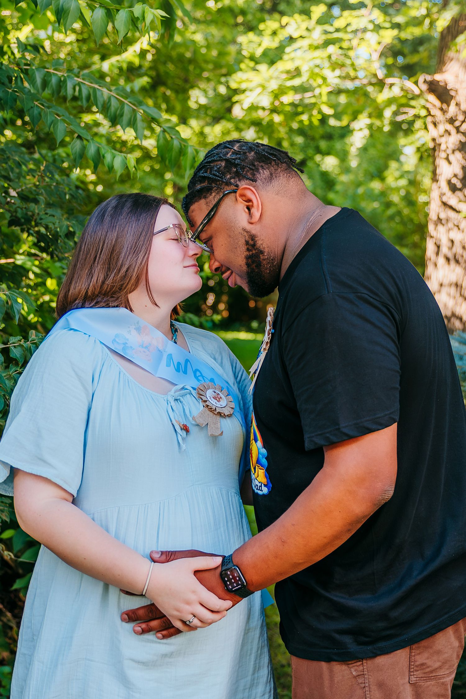 Expecting couple sharing a tender moment outdoors, touching baby bump, surrounded by lush green trees.
