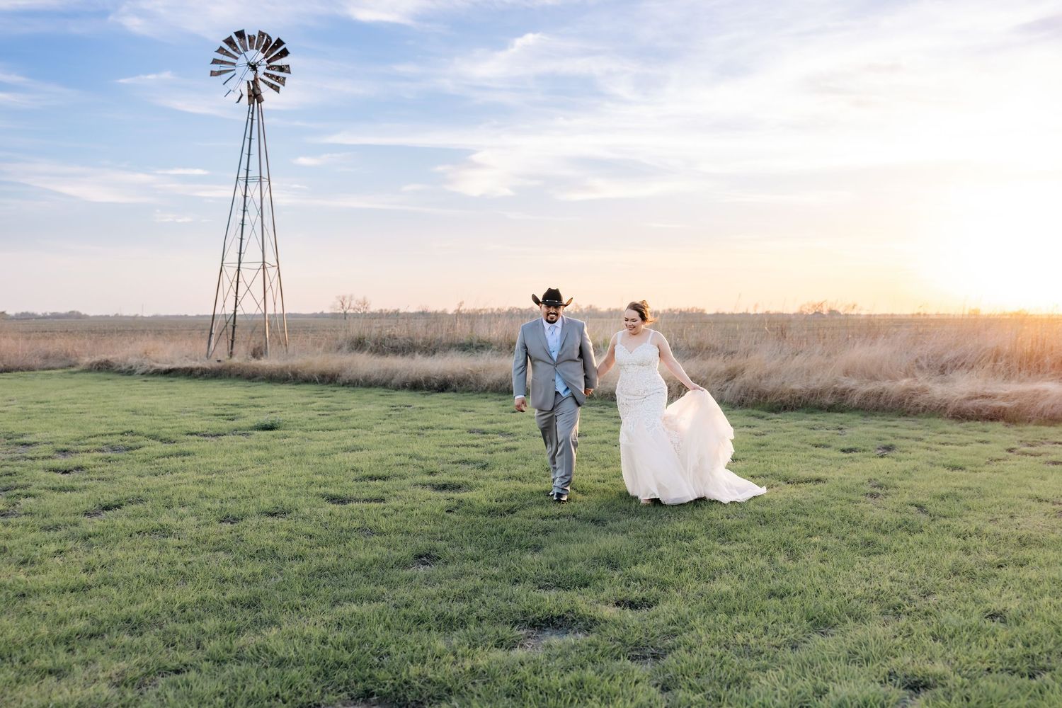Bride and groom walk hand in hand through a green field at golden sunset with a windmill in the distance.