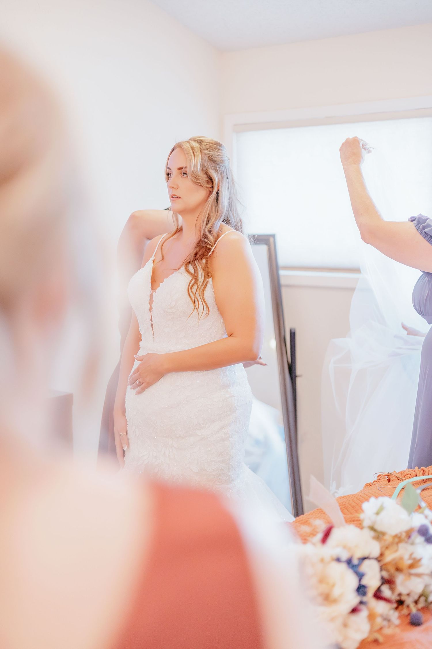 Blonde bride in lace wedding gown being helped with veil by bridesmaid in bright dressing room.