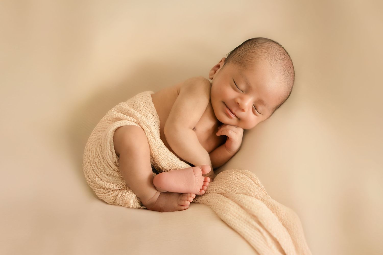 Peaceful newborn baby sleeping curled up on a soft beige blanket, smiling in a cozy cream wrap.